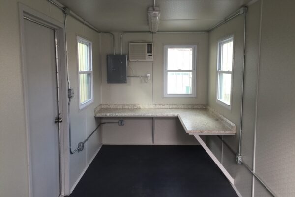 A complete interior view of a modified shipping/storage container that has finished white walls, overhead lighting, a brown and white multi-colored countertop, and electrical panel and outlets.