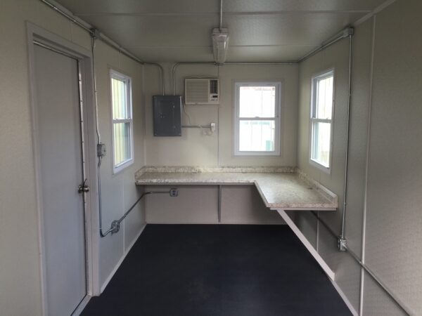 A complete interior view of a modified shipping/storage container that has finished white walls, overhead lighting, a brown and white multi-colored countertop, and electrical panel and outlets.